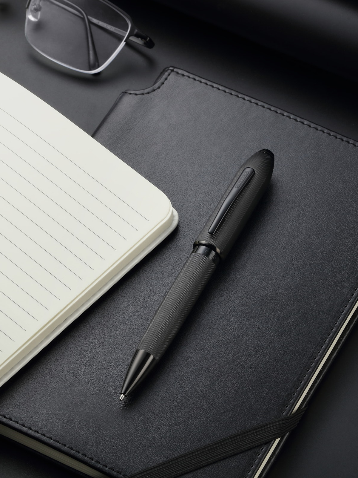 Black pen on a black leather notebook with a white lined page, glasses in the background.