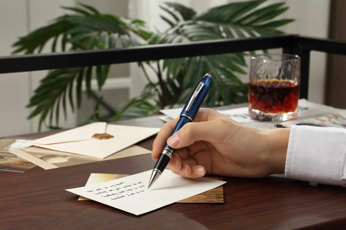 Hand holding a pen over a letter with a glass of dark liquid on a table, surrounded by plants.