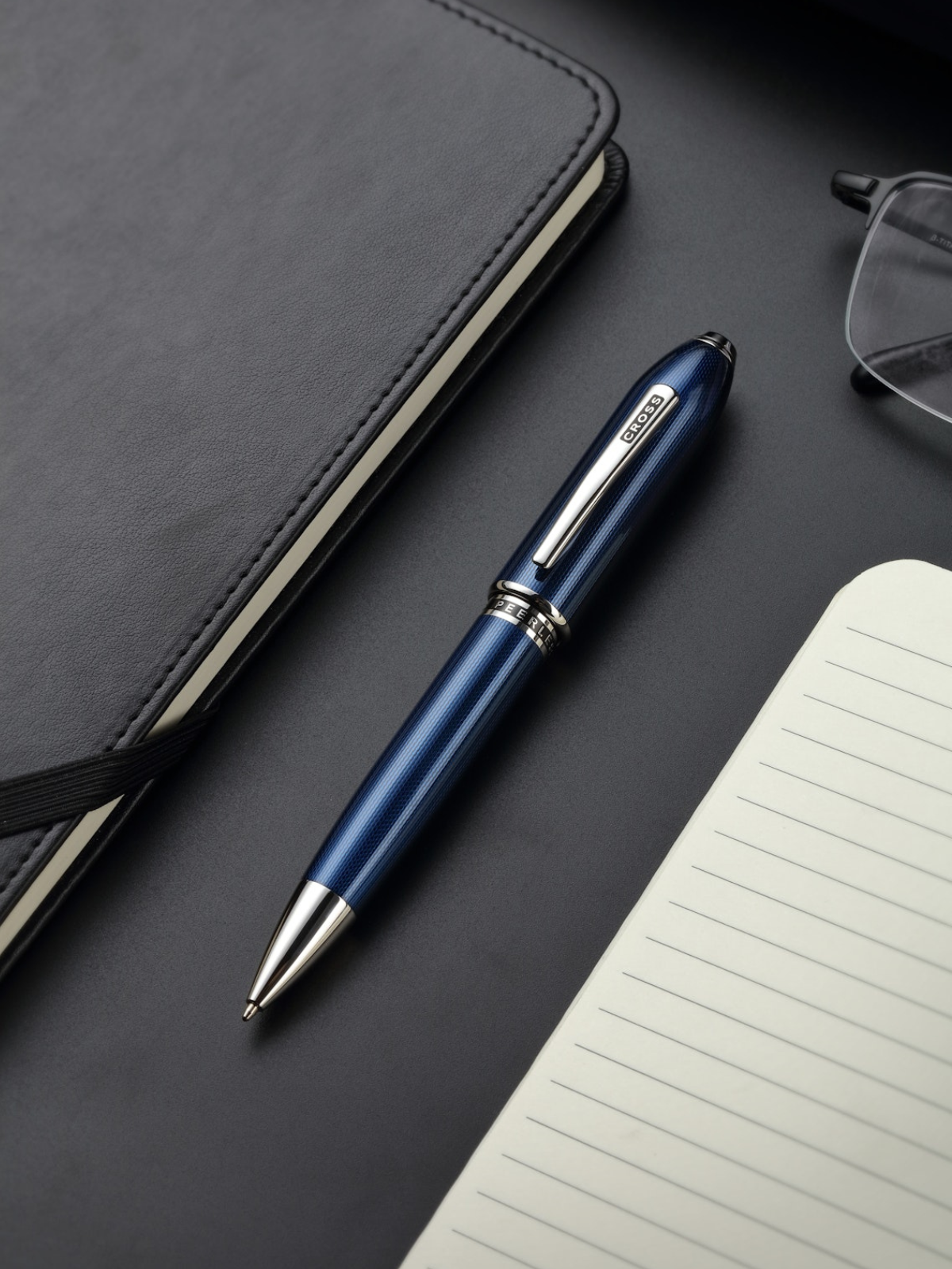 Blue pen with silver accents on a dark surface with a notebook and glasses.